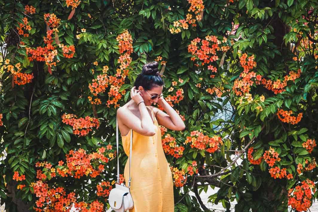 Girl in yellow dress standing in front of a wall of orange flowers and green leaves, enjoying the beauty of nature.